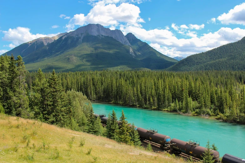 a train traveling through a lush green forest next to a river, by Brigette Barrager, pexels contest winner, banff national park, behind that turquoise mountains, green gas spreading across land, piping