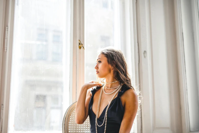 a woman in a black dress sitting in front of a window, by Julia Pishtar, pexels contest winner, gold and pearl necklaces, pearl choker, sydney hanson, pearls and gold chains