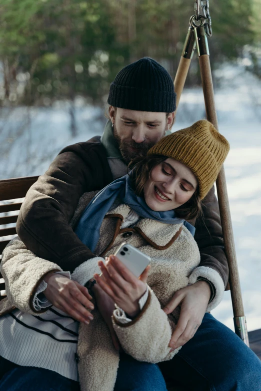 a man and woman sitting on a swing in the snow, by Eero Järnefelt, trending on pexels, she is holding a smartphone, beanie, cinema still, hug