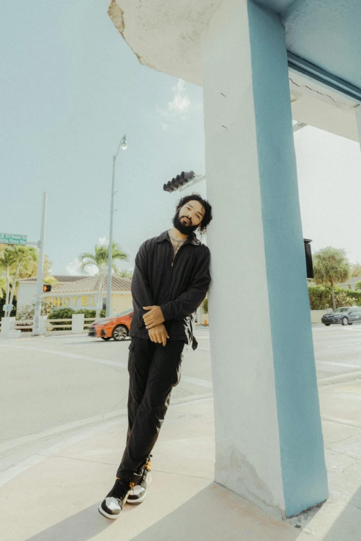 a man standing on the side of a building, standing in a parking lot, black curly beard, with palm trees in the back, laying down