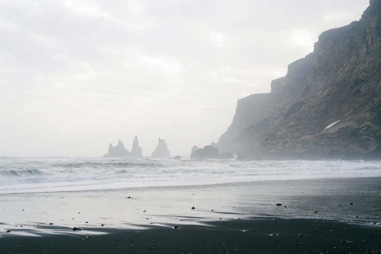 a person standing on a beach with a surfboard, by Carey Morris, pexels contest winner, tall stone spires, gray fog, black sand, coastal cliffs