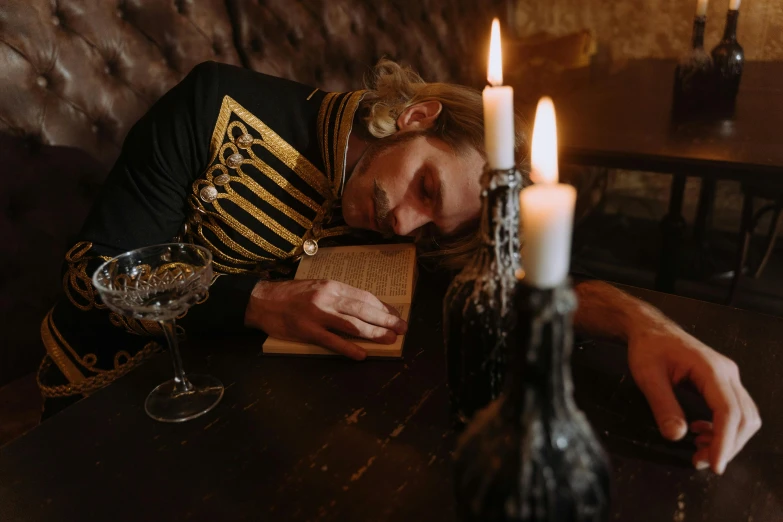 a man laying on top of a wooden table next to a lit candle, an album cover, inspired by Vasily Perov, pexels contest winner, renaissance, xix century military outfit, drinking, cinematic outfit photo, holding grimoire