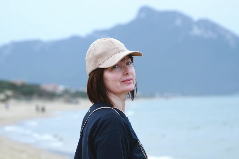 a woman standing on a beach next to the ocean, inspired by Zhang Kechun, renaissance, baseball cap, beige mist, plain, official product photo