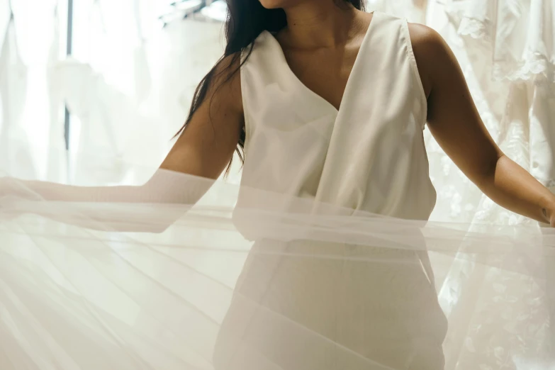 a woman standing in front of a rack of wedding dresses, trending on pexels, light and space, white sarong, aerial silk, wearing translucent sheet, wearing white v - neck top