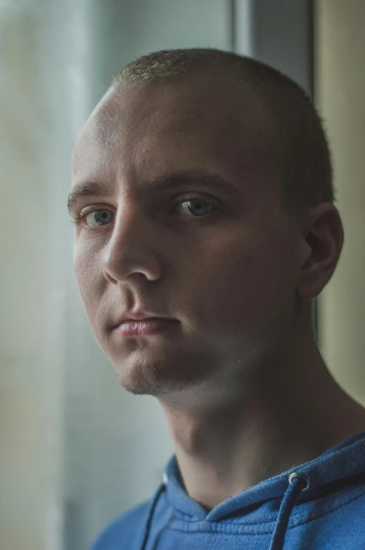 a man in a blue hoodie looking out a window, a portrait, inspired by Alexey Venetsianov, unsplash, hyperrealism, shaven, looking directly at the camera, transgender, no hair