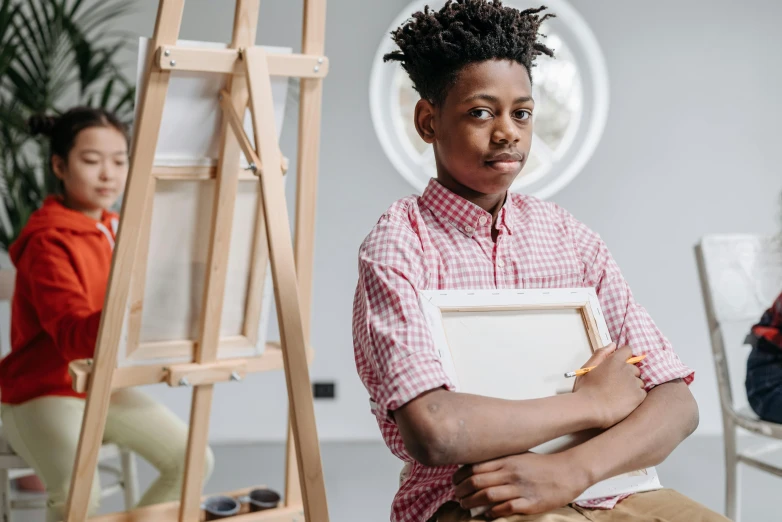 a young boy sitting on a chair in front of a easel, pexels contest winner, academic art, black teenage boy, androgynous male, holding a clipboard, over his shoulder