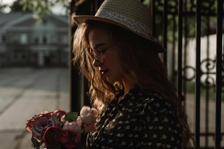 a woman standing in front of a gate holding a bouquet of flowers, pexels contest winner, romanticism, wearing a fedora, profile image, girl with glasses, morning hard light