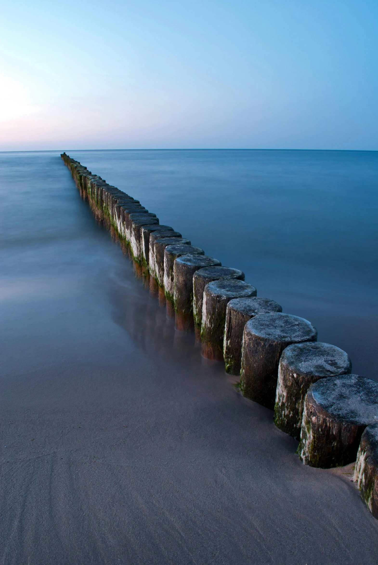 a long exposure photo of a pier in the middle of the ocean, by Karl Walser, wooden logs, in a row, evening light, sand