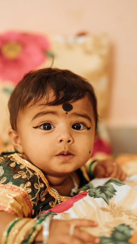 a close up of a baby laying on a bed, pexels contest winner, hyperrealism, dressed in a sari, proud serious expression, square, high quality photo