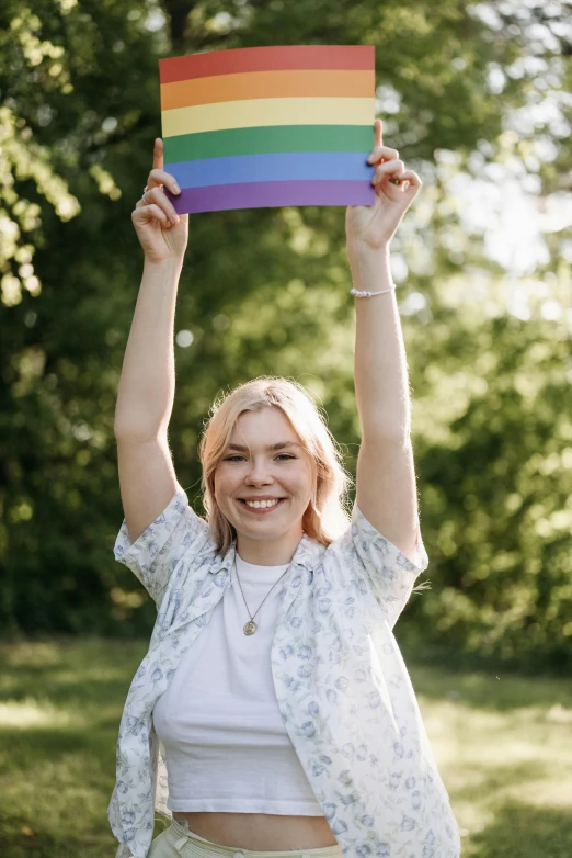 a woman holding up a rainbow flag in a park, by Alice Mason, portrait of anya taylor-joy, woman his holding a sign, pose(arms up + happy), a blond