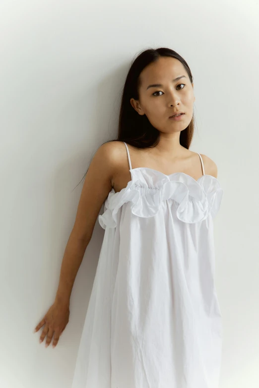 a woman in a white dress leaning against a wall, inspired by Cui Bai, unsplash, gutai group, sleepwear, ruffles, wearing a camisole, full product shot