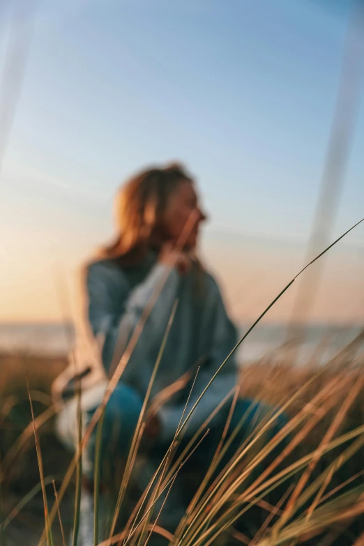 a woman sitting on top of a beach next to tall grass, a picture, trending on unsplash, happening, golden hour closeup photo, field - blur, relaxed. blue background, looking out a window