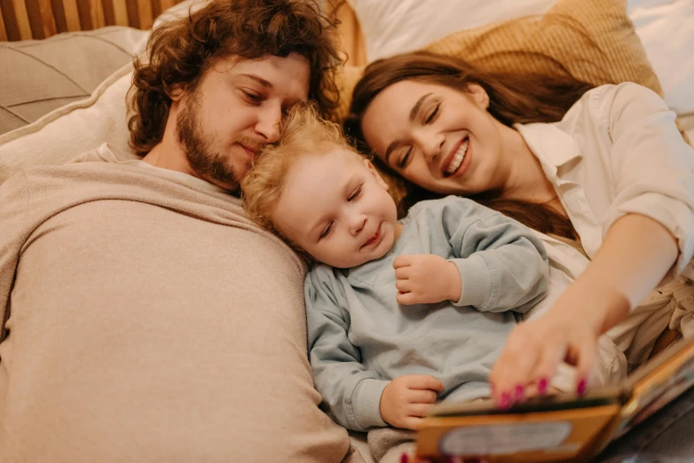 a man and woman laying in bed with a baby, a cartoon, pexels, incoherents, curled up on a book, happy kid, profile image, medium close-up shot