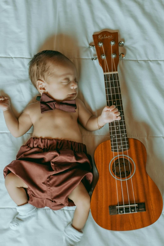 a baby laying on top of a bed next to a guitar, by Nina Hamnett, pexels contest winner, symbolism, ukulele, bows, maroon, feels good man