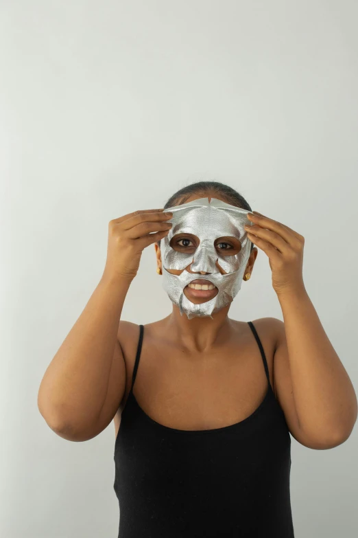 a woman putting a mask on her face, white metallic, with brown skin, front facing, dramatic product shot