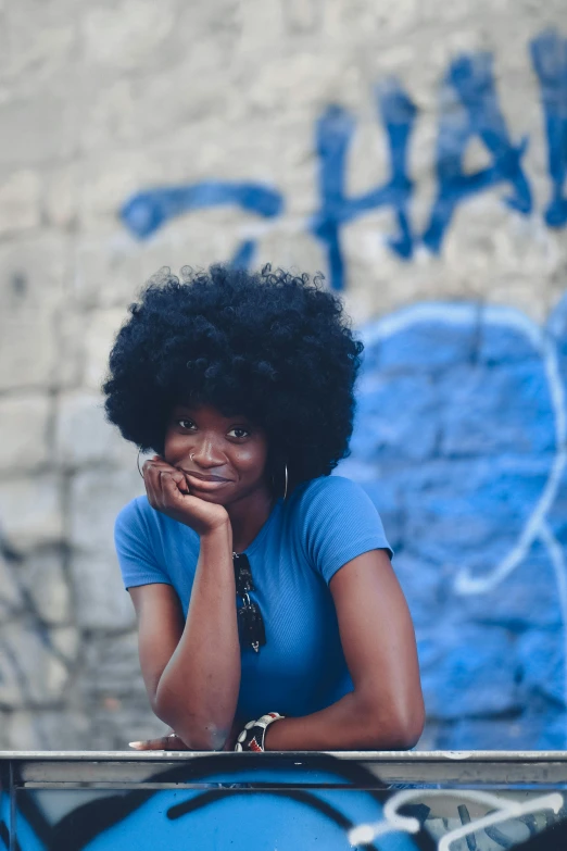 a woman sitting in front of a graffiti covered wall, an album cover, pexels contest winner, afrofuturism, natural hair, ((blue)), blacks and blues, handsome girl