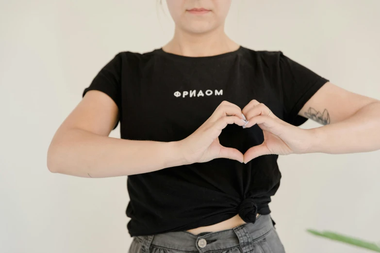 a woman making a heart with her hands, by Nina Hamnett, trending on pexels, black t shirt, ukrainian, omoide emanon, logo