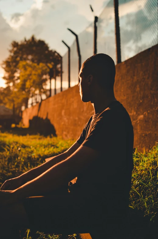 a man sitting on the ground in front of a fence, looking off into the sunset, profile image