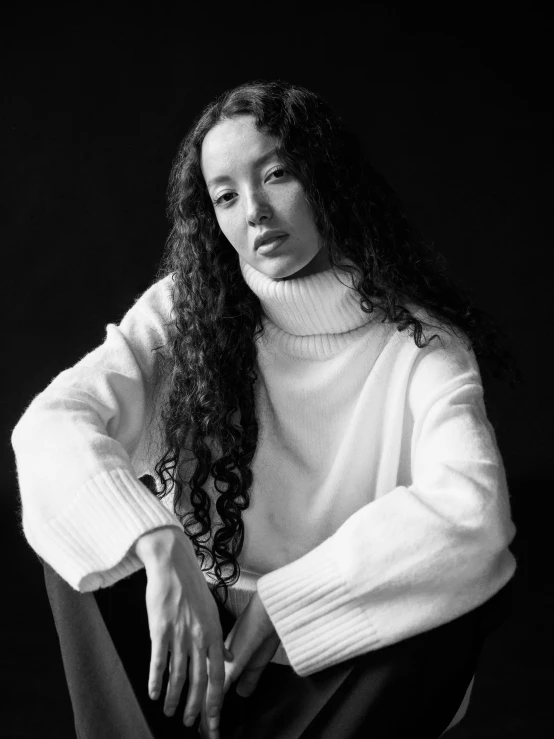 a black and white photo of a woman sitting on a chair, a black and white photo, unsplash contest winner, realism, in white turtleneck shirt, bella poarch, with long curly, wearing a sweater