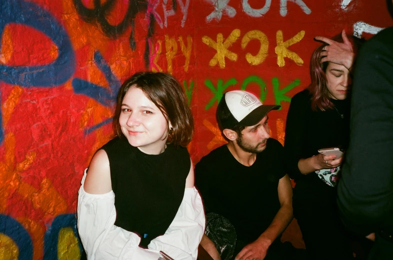 a group of people standing in front of a wall covered in graffiti, an album cover, inspired by Elsa Bleda, pexels contest winner, antipodeans, joey king, smug smile, profile image, drunk