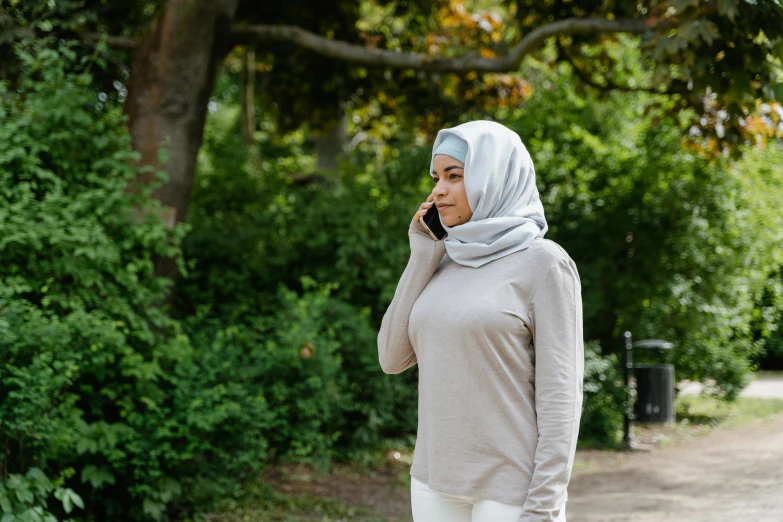 a woman in a hijab talking on a cell phone, light grey, in a park, grey and silver, beige