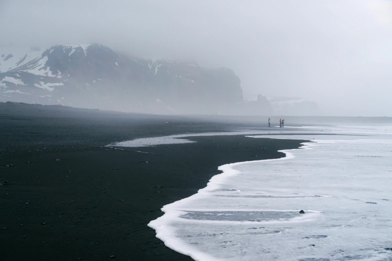a man standing on top of a beach next to the ocean, pexels contest winner, romanticism, iceland, people walking in the distance, black fog, in an icy river
