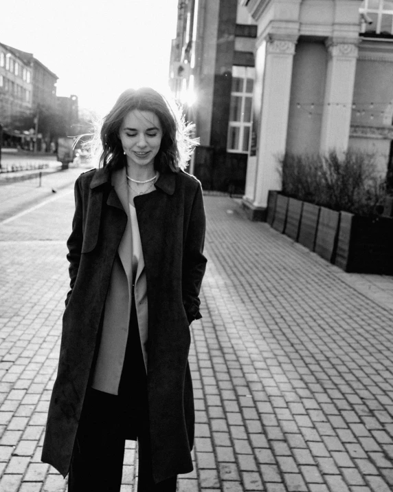 a black and white photo of a woman standing on a sidewalk, emilia clarke, trench coat, morning glow, moleksandra shchaslyva