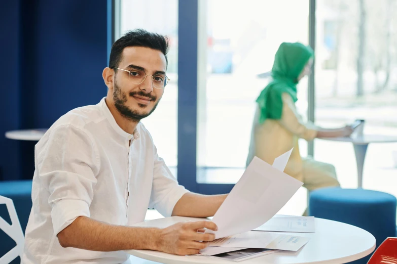 a man sitting at a table with papers in front of him, by Ismail Acar, pexels contest winner, subject is smiling, an arab standing watching over, lachlan bailey, commercial