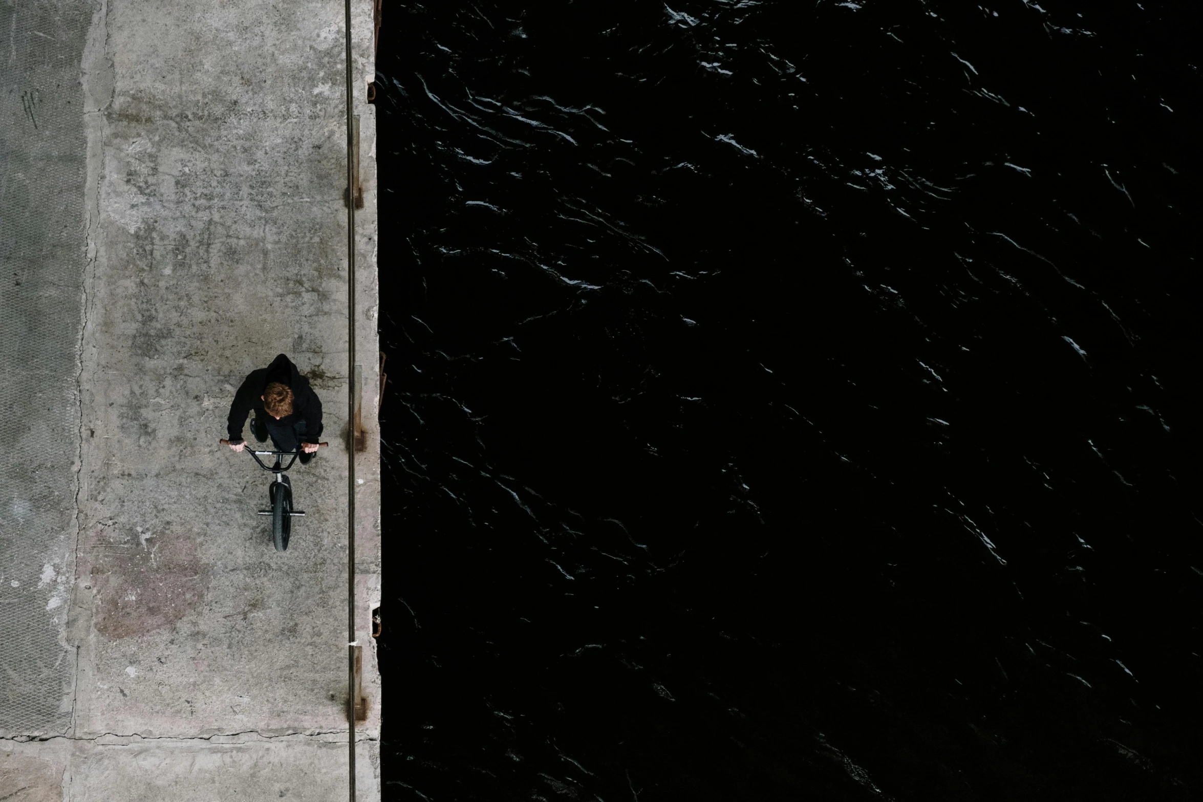 a person sitting on a ledge next to a body of water, an album cover, inspired by Jan Rustem, pexels contest winner, bicycle, aerial shot from the drone, on a black wall, harbor