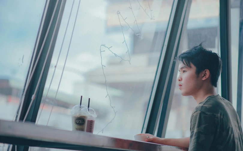 a boy sitting at a table looking out a window, inspired by Liam Wong, trending on unsplash, realism, young asian woman, drinking boba tea, scene from live action movie, proud looking away