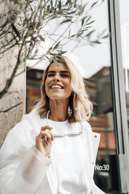 a woman standing next to a tree in front of a building, by Sara Saftleven, trending on unsplash, big smile white teeth, wearing white robes, exiting store, white highlights in hair