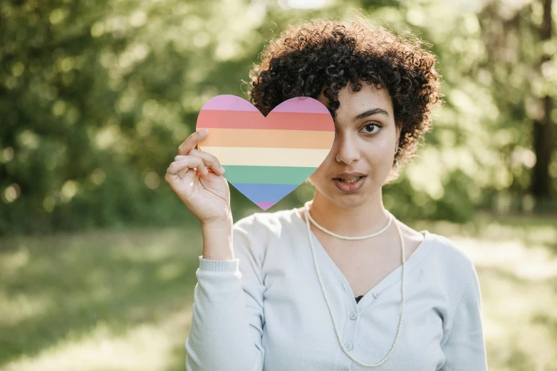 a woman holding a rainbow heart in front of her face, trending on pexels, curly pixie cut hair, holding up a large shield, 🚿🗝📝, young male