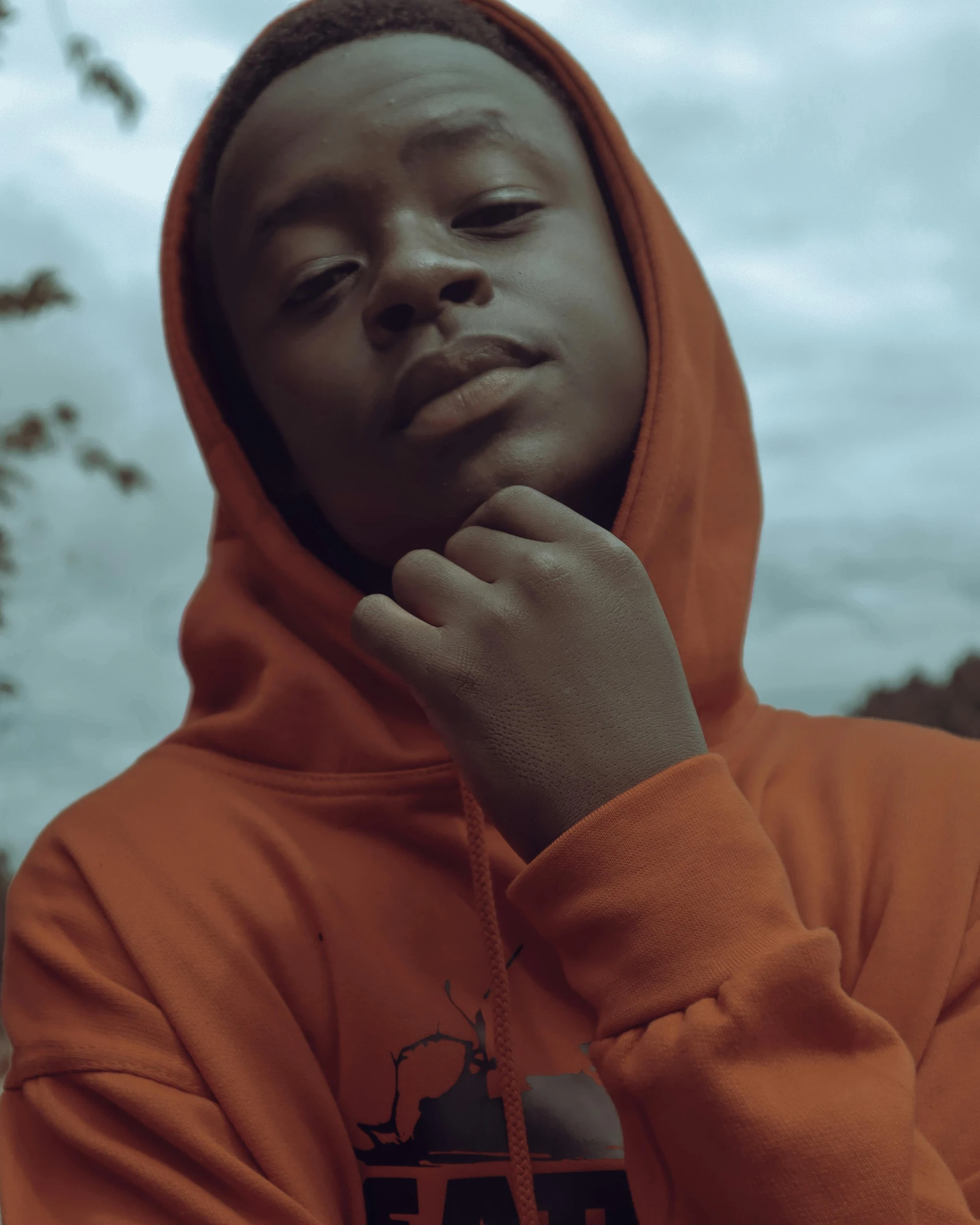 a man in an orange hoodie poses for a picture, an album cover, inspired by Xanthus Russell Smith, trending on unsplash, black teenage boy, lgbtq, still frame from a movie, grainy footage