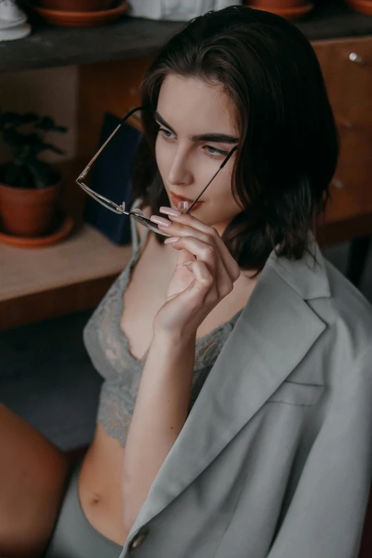 a woman sitting on a chair holding a pair of glasses, inspired by Elsa Bleda, trending on pexels, sexy lips :5 stylish, grey robe, bralette, wearing a worn out suit