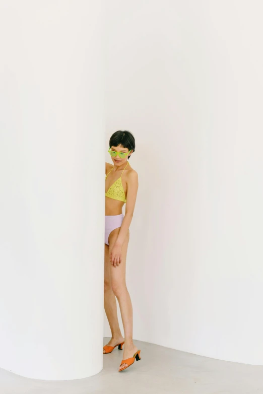 a woman standing in front of a white wall, inspired by Ren Hang, yellow purple green, underwear ad, masked person in corner, rinko kawauchi