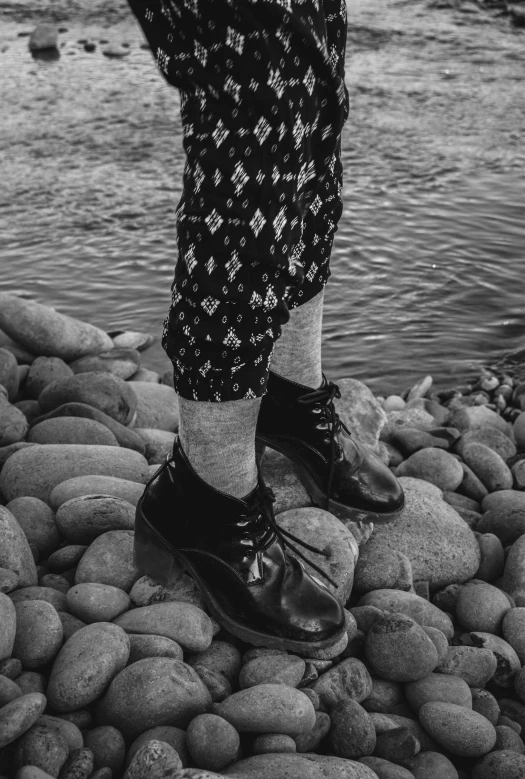 a black and white photo of a person standing on rocks, a black and white photo, unsplash, wearing skirt and high socks, patterned clothing, duck shoes, ((rocks))