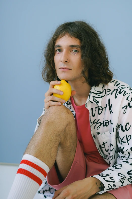 a man sitting on a couch holding an orange, an album cover, inspired by Davide Sasselli, wearing a lemon, holding daisy, looking away from camera, wearing casual clothing