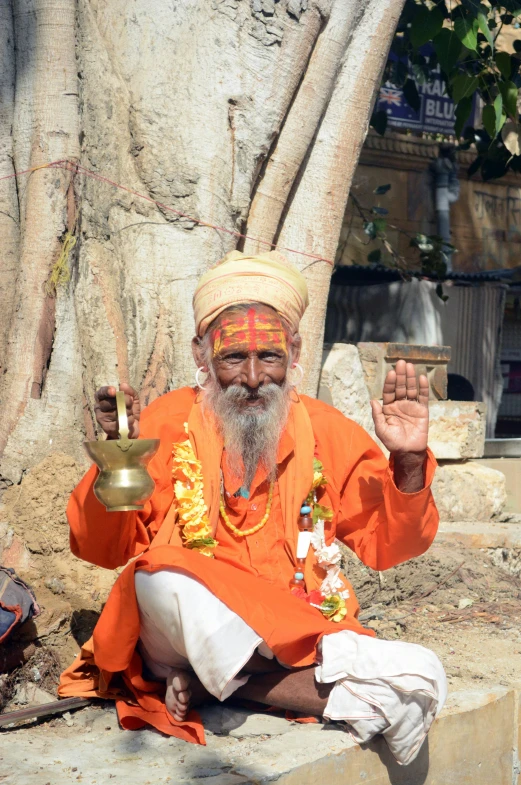 a man sitting on a ledge next to a tree, a statue, samikshavad, orange and yellow costume, blessing hands, an oldman, facing the camera