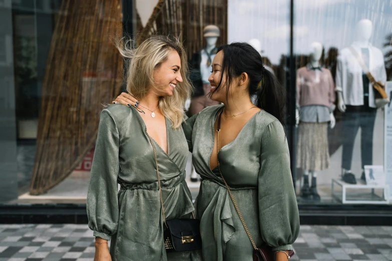 two women standing next to each other in front of a store, by Emma Andijewska, trending on pexels, happening, dressed in a green robe, jumpsuit, slightly tanned, wearing a cocktail dress