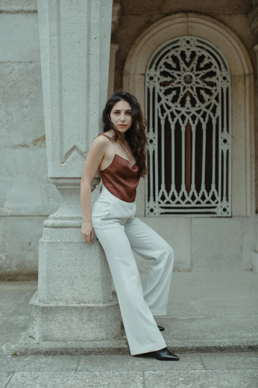 a woman sitting on a pillar in front of a building, inspired by Luis Paret y Alcazar, trending on pexels, baroque, wearing a camisole, brown pants, promotional image, wearing off - white style