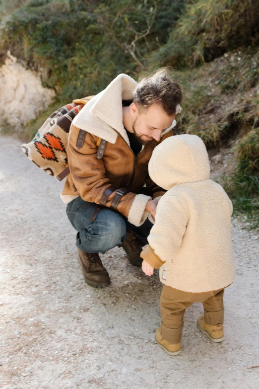 a man kneeling next to a small child on a dirt road, pexels contest winner, wearing a brown leather coat, cuddling, avatar image, beige