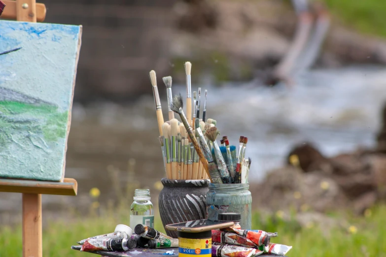 a table topped with art supplies next to a river, pexels contest winner, plein air, fan favorite, holding paintbrushes, in the world of andrew wyeth, panoramic shot