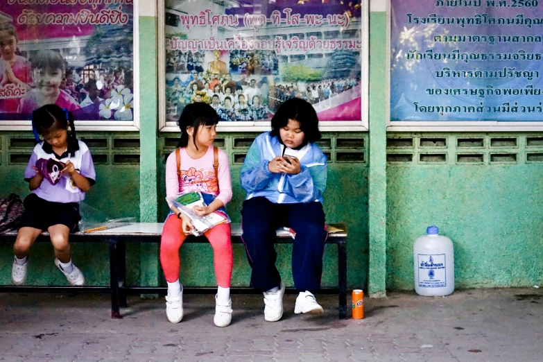 three girls sitting on a bench looking at their cell phones, a photo, by Nathalie Rattner, thailand, square, bus stop, a book