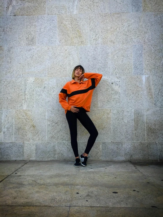 a woman in an orange jacket leaning against a wall, by Robbie Trevino, wearing track and field suit, orange and black tones, various pose, trending on r/techwearclothing