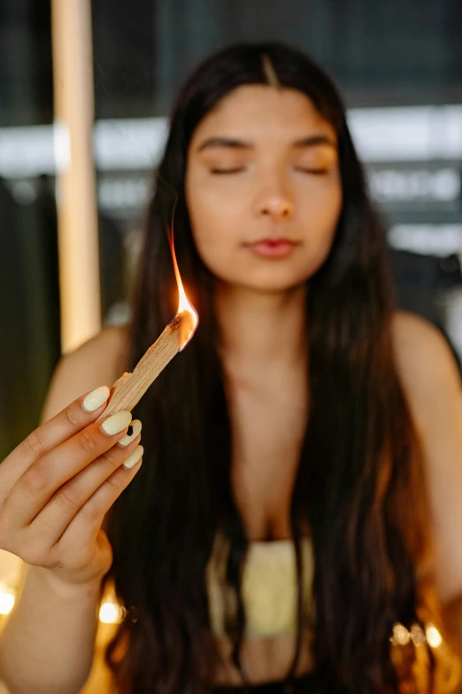 a woman sitting at a table with a candle in her hand, trending on pexels, smoking a blunt, female with long black hair, holding a burning wood piece, light tan