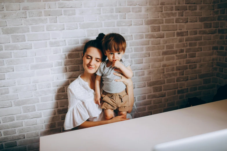 a woman holding a small child in her arms, pexels contest winner, sitting on a desk, avatar image, leaning on the wall, monitor