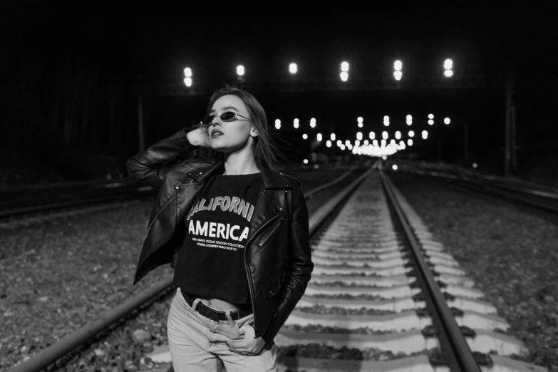 a woman standing on a train track at night, a black and white photo, unsplash, american realism, black leather jacket, american progress, woman in streetwear, california;
