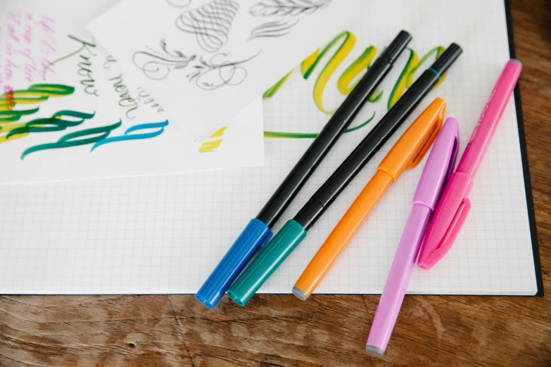 a bunch of pens sitting on top of a notebook, colouring - in sheet, brand colours are green and blue, flowing lettering, product shot