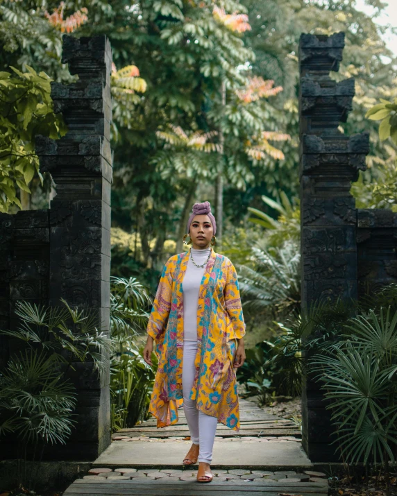 a woman standing on a wooden walkway in a garden, an album cover, by Lily Delissa Joseph, sumatraism, wearing rainbow kimono, promo image, batik, photo for vogue