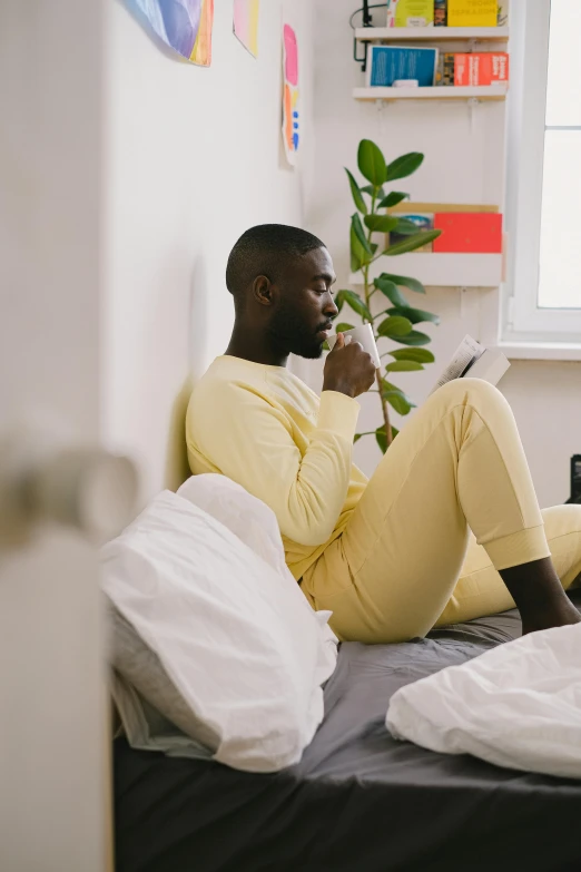 a man sitting on a bed reading a book, a cartoon, trending on pexels, yellow clothes, black man, drinking a coffee, wearing a track suit